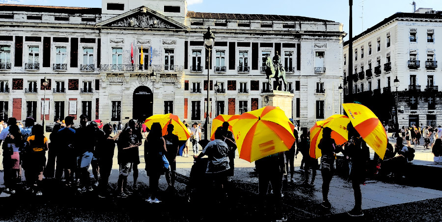 Sombrillas amarillas bajo el sol en una plaza, la gente aparece al estilo sombras chinas